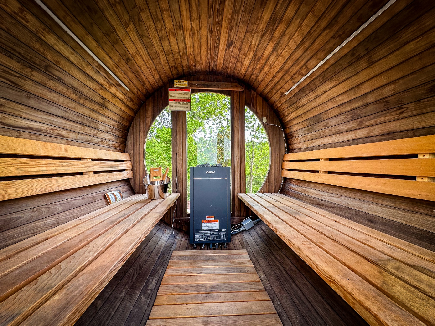 Interior of the cedar barrel sauna with tiered wooden benches, Harvia heater, and a round window framing green trees