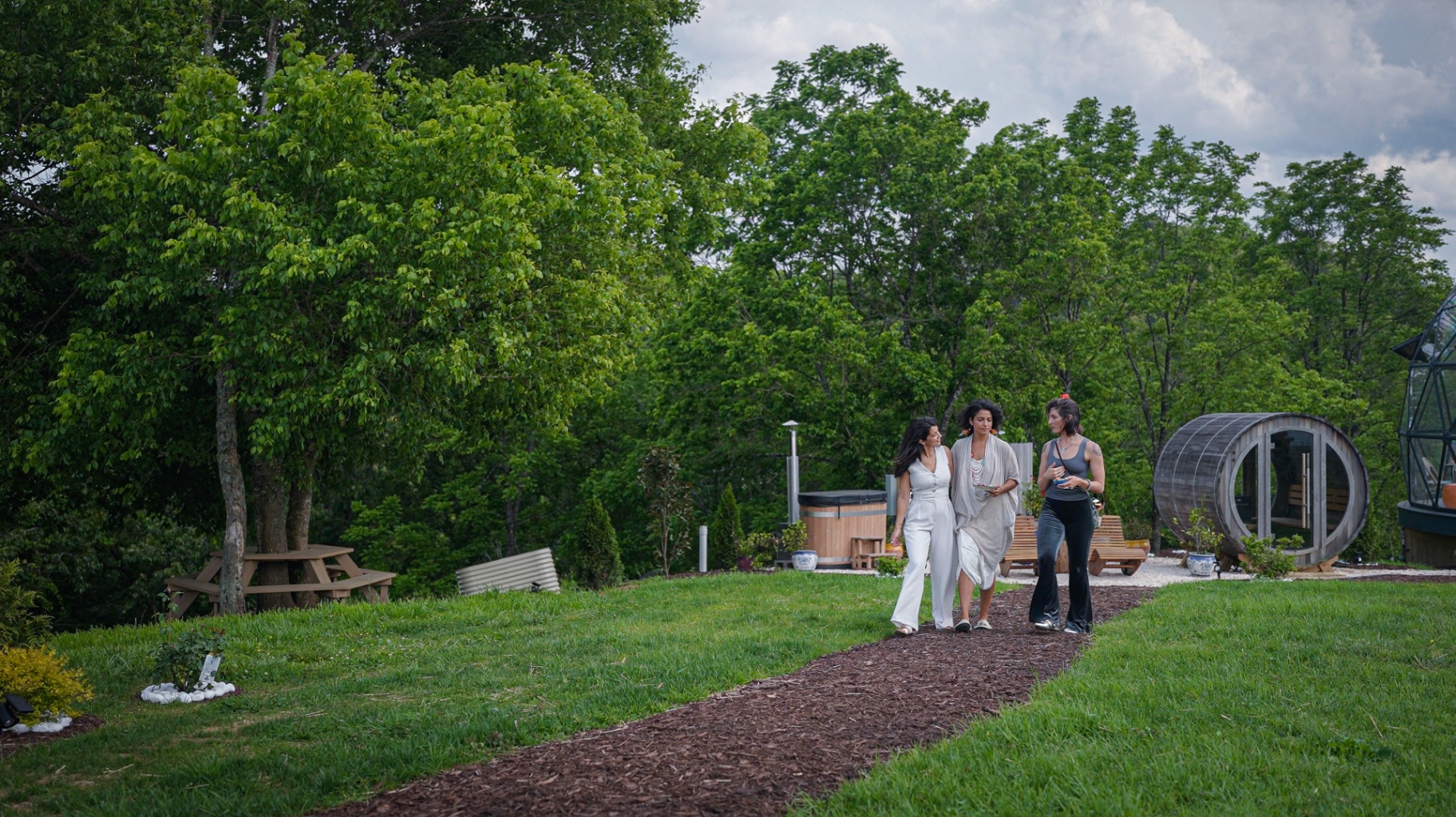 Three women walking the path together past the cedar sauna, mid-conversation