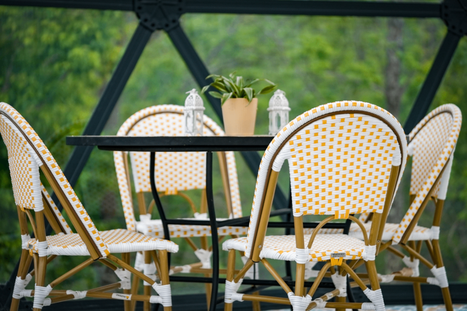 Yellow-and-white rattan bistro chairs around a black table with white Moroccan lanterns and a small plant, greenery blurred beyond the dome frame