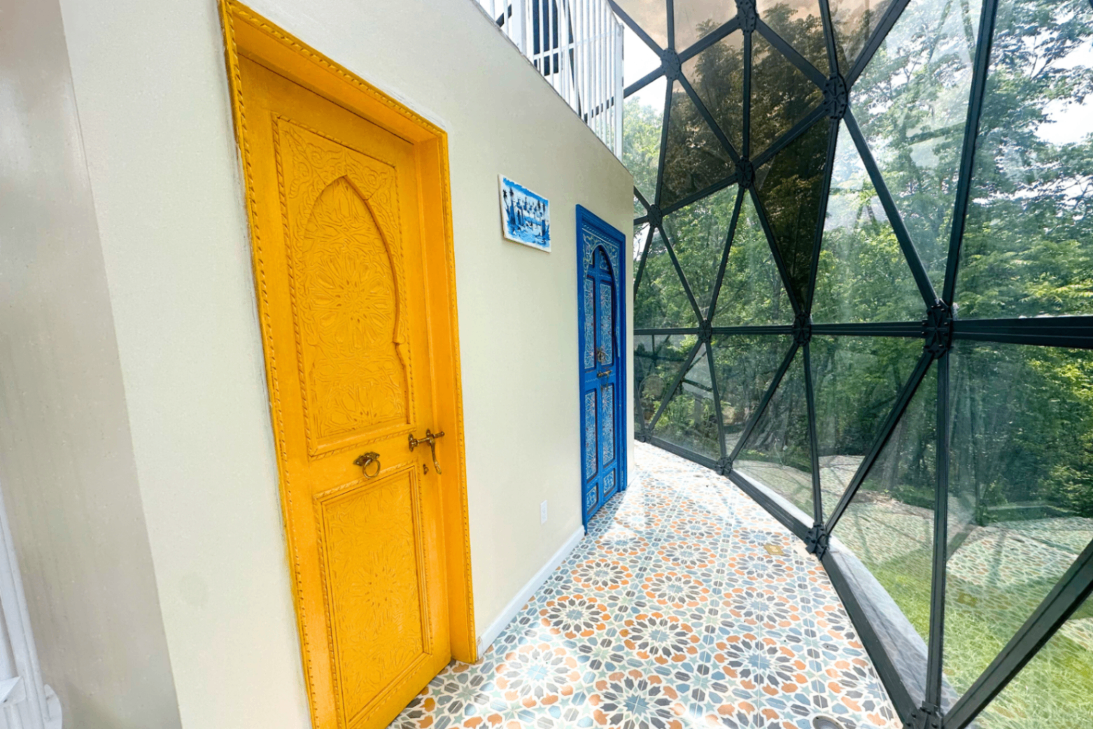 Tiled corridor with a carved saffron-yellow door, a cobalt-blue door beyond, and a geometric star-pattern floor running to the geodesic glass