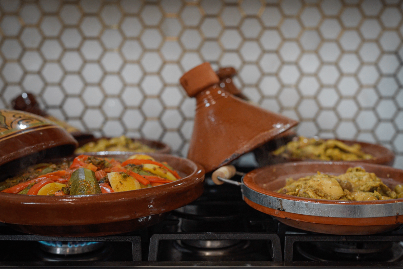 Three clay tagines simmering on the stove, one lid lifted to show vegetables and saffron chicken