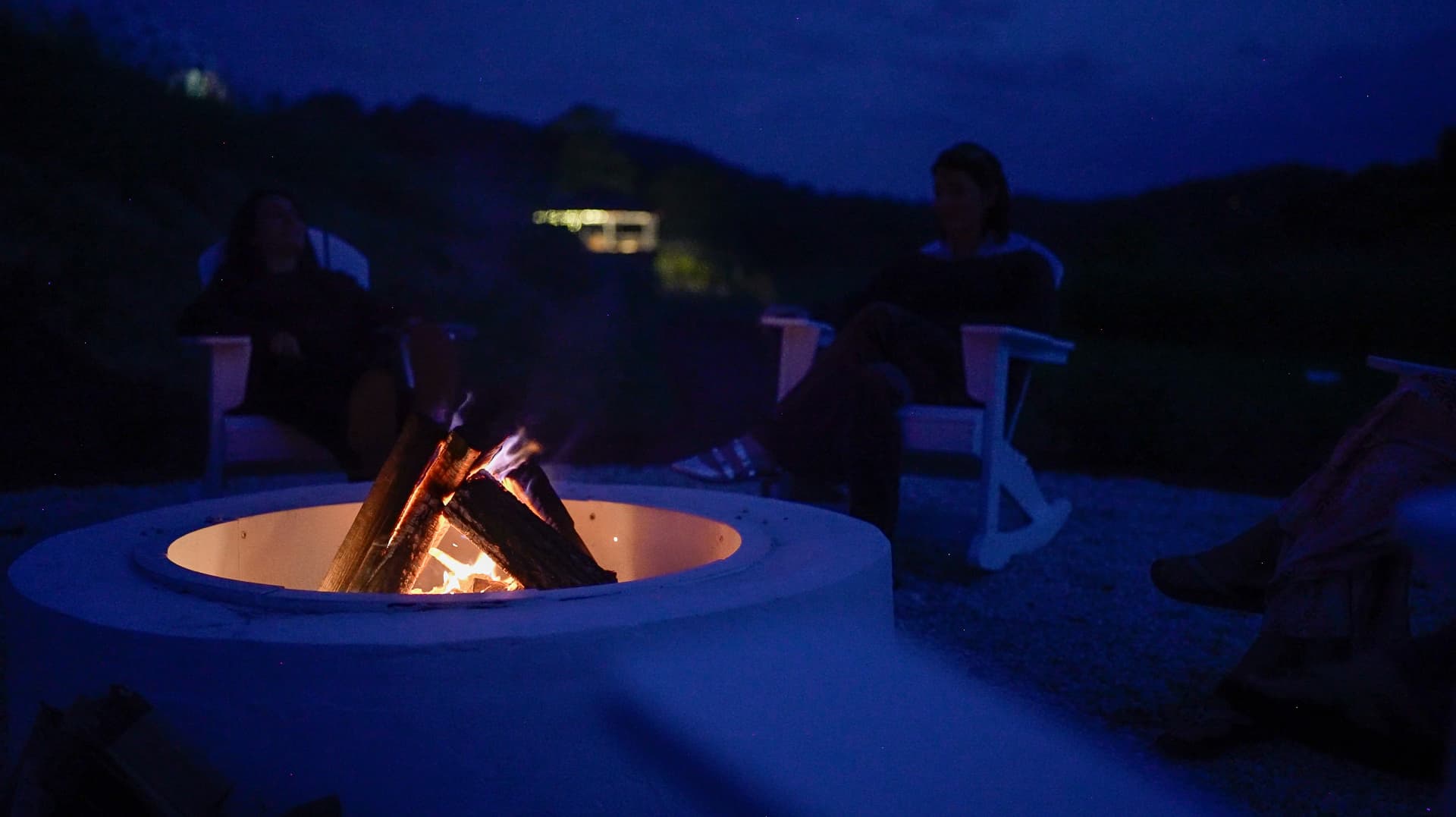 Two guests seated by the lit fire pit after dark, with the hills behind them