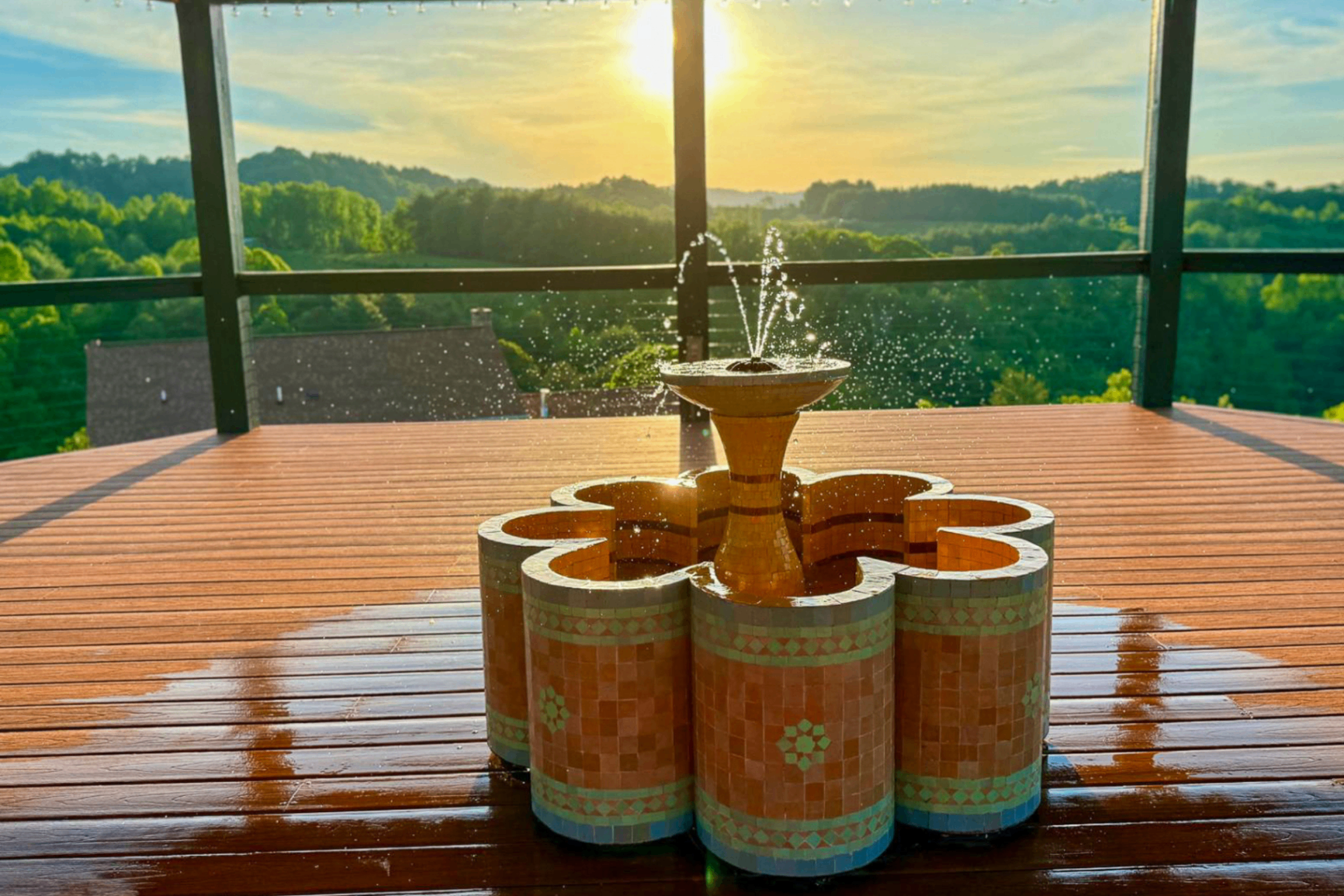 Flower-shaped tiled mosaic fountain on the pavilion deck with water arcing at golden hour, the valley behind