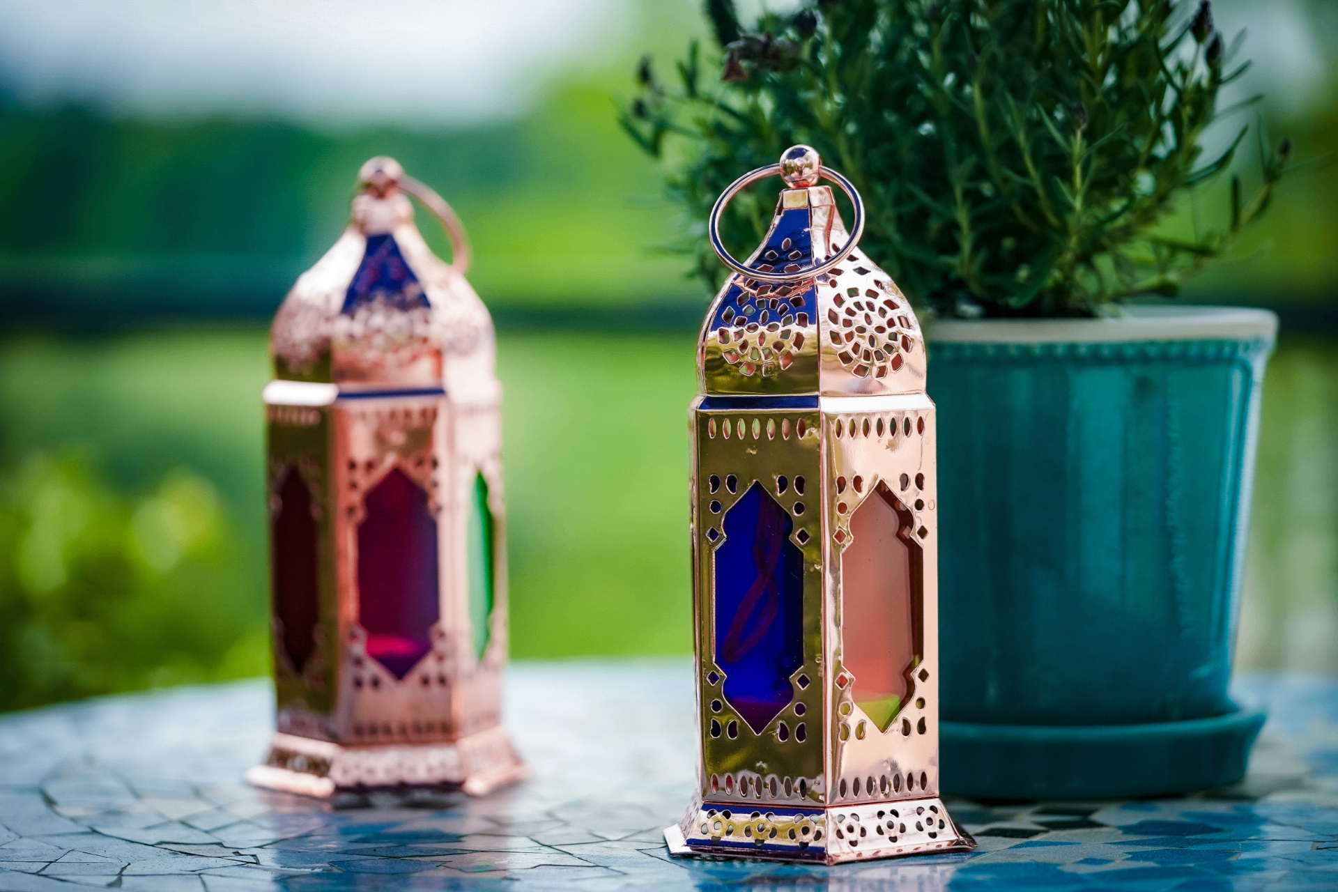 Two pierced copper Moroccan lanterns with stained glass panels beside a rosemary plant on a blue mosaic table