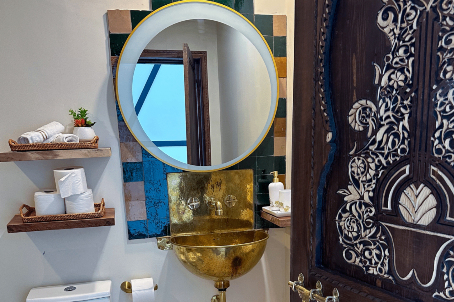 Hammered gold sink beneath a round mirror, framed by hand-glazed zellige tile in green, blue, and terracotta, with a carved wooden door at right