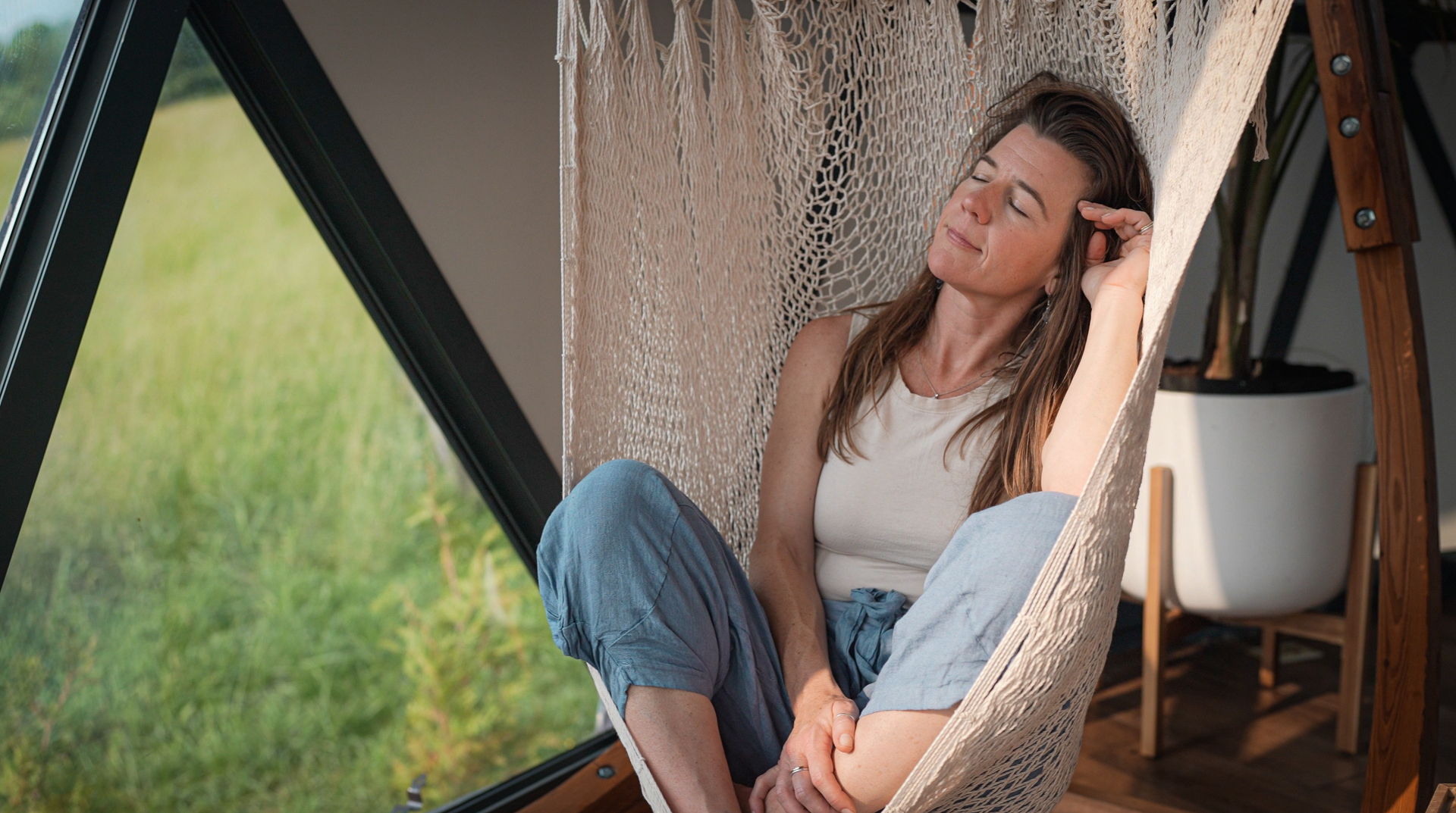 Someone resting in a hammock chair inside a geodesic dome, eyes closed, the green field beyond the glass